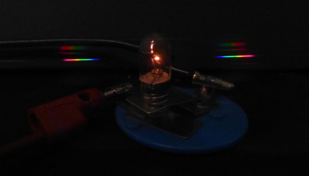 A photograph of an incandescent bulb viewed through a diffraction grating. The bulb itself is barely visible except for a yellow/white glow where the filament is. To each side (left and right) of the bulb can be seen a rainbow of light produced by the diffraction grating.