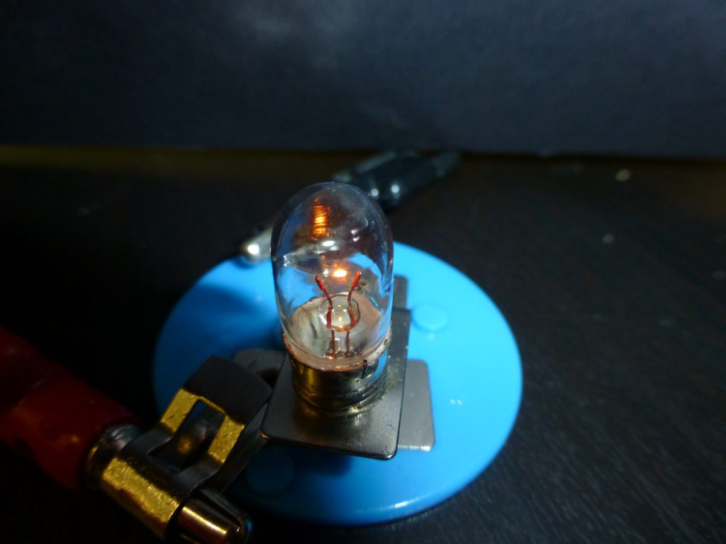 A photograph of a small bulb. The surroundings are very dark. Visible is a glass bulb containing some wires connecting to a glowing filament. The filament is glowing yellow/white in color.