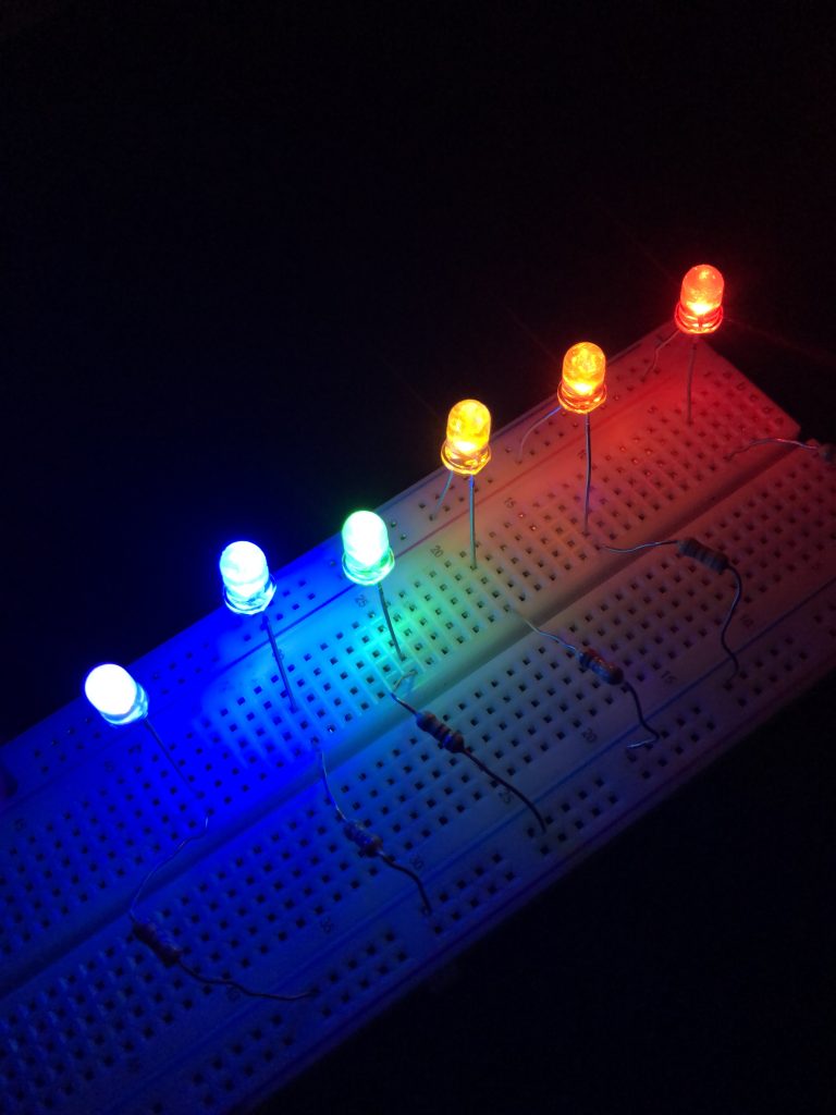 A photograph of six LEDs placed in a breadboard, all lit up and emitting light. (They are in a dark room to provide contrast with the background.) The individual LED colors are red, orange, yellow, green, blue, and violet.