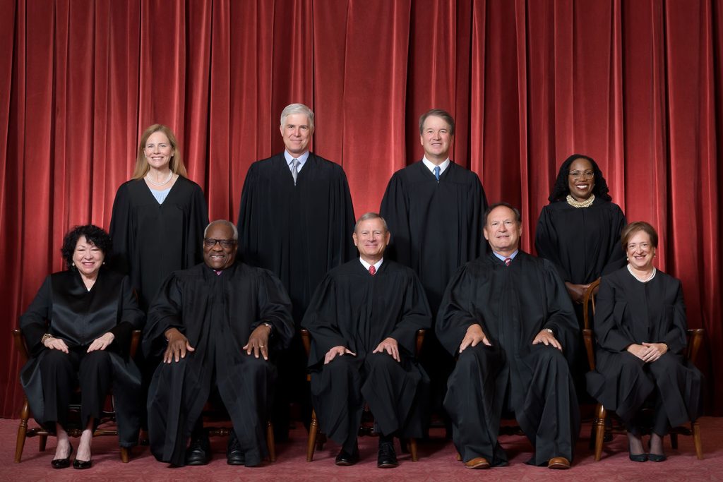 Formal group photograph of the Supreme Court as it was comprised on June 30, 2022. The Justices are posed in front of red velvet drapes and arranged by seniority, with five seated and four standing. Seated from left are Justices Sonia Sotomayor, Clarence Thomas, Chief Justice John G. Roberts, Jr., and Justices Samuel A. Alito and Elena Kagan. Standing from left are Justices Amy Coney Barrett, Neil M. Gorsuch, Brett M. Kavanaugh, and Ketanji Brown Jackson.