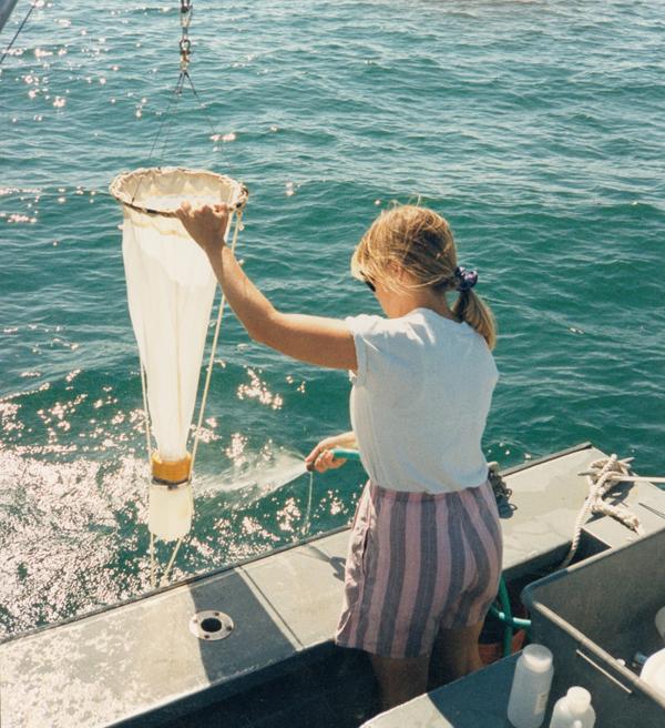 A woman in a boat holds a plankton net above the water.