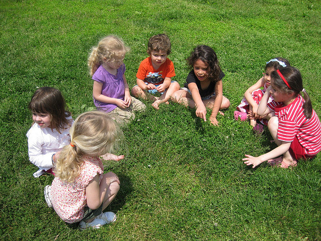 Color photograph of young children sitting in a circle playing a game or looking at something in the grass