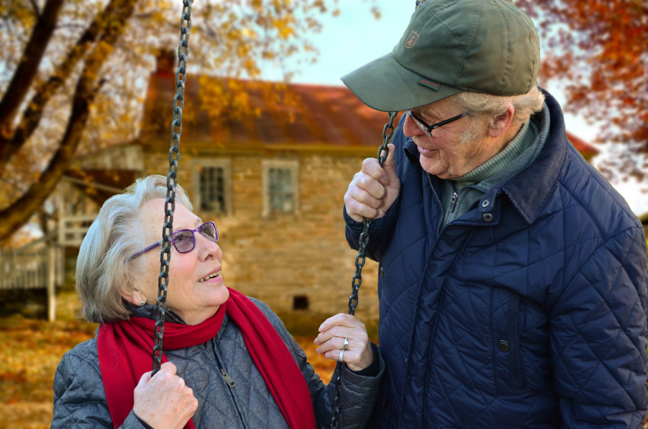 Color photogrpahy of an elderly woman on a swing and an elderly man looking down at her. It is fall.